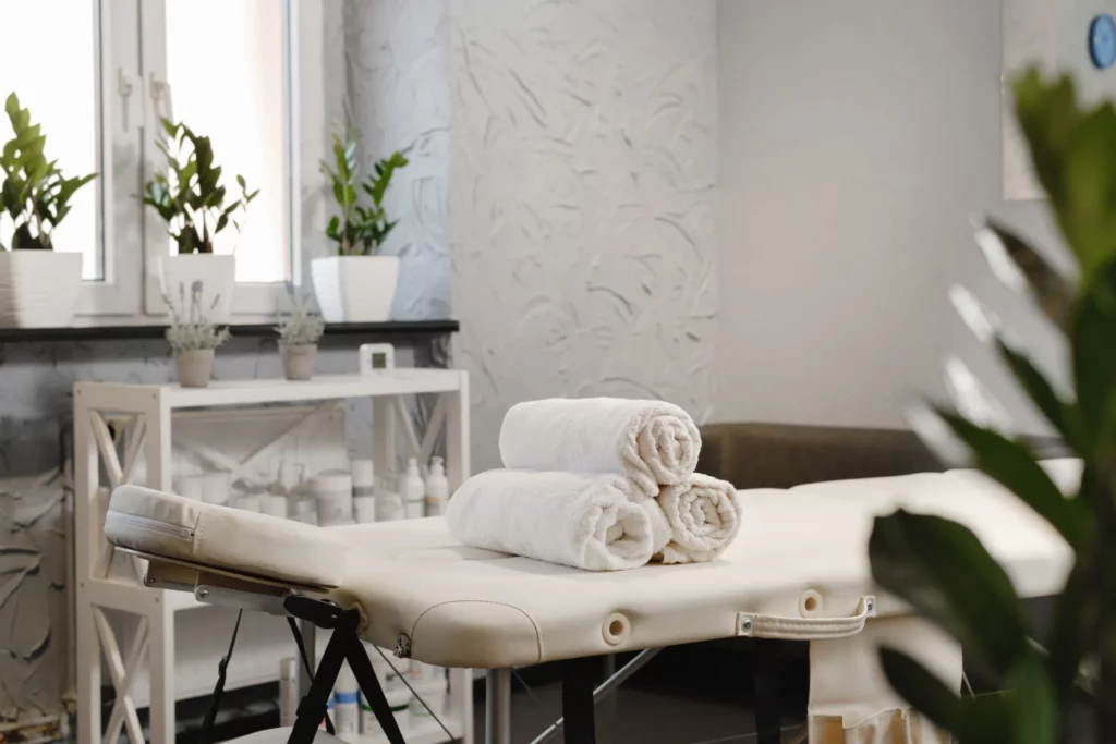 A white massage table with three white rolled towels placed atop it in a white massage room. Green plants in white planters sit at the windowsill, and a white shelving rack with massage products faces the massage table.