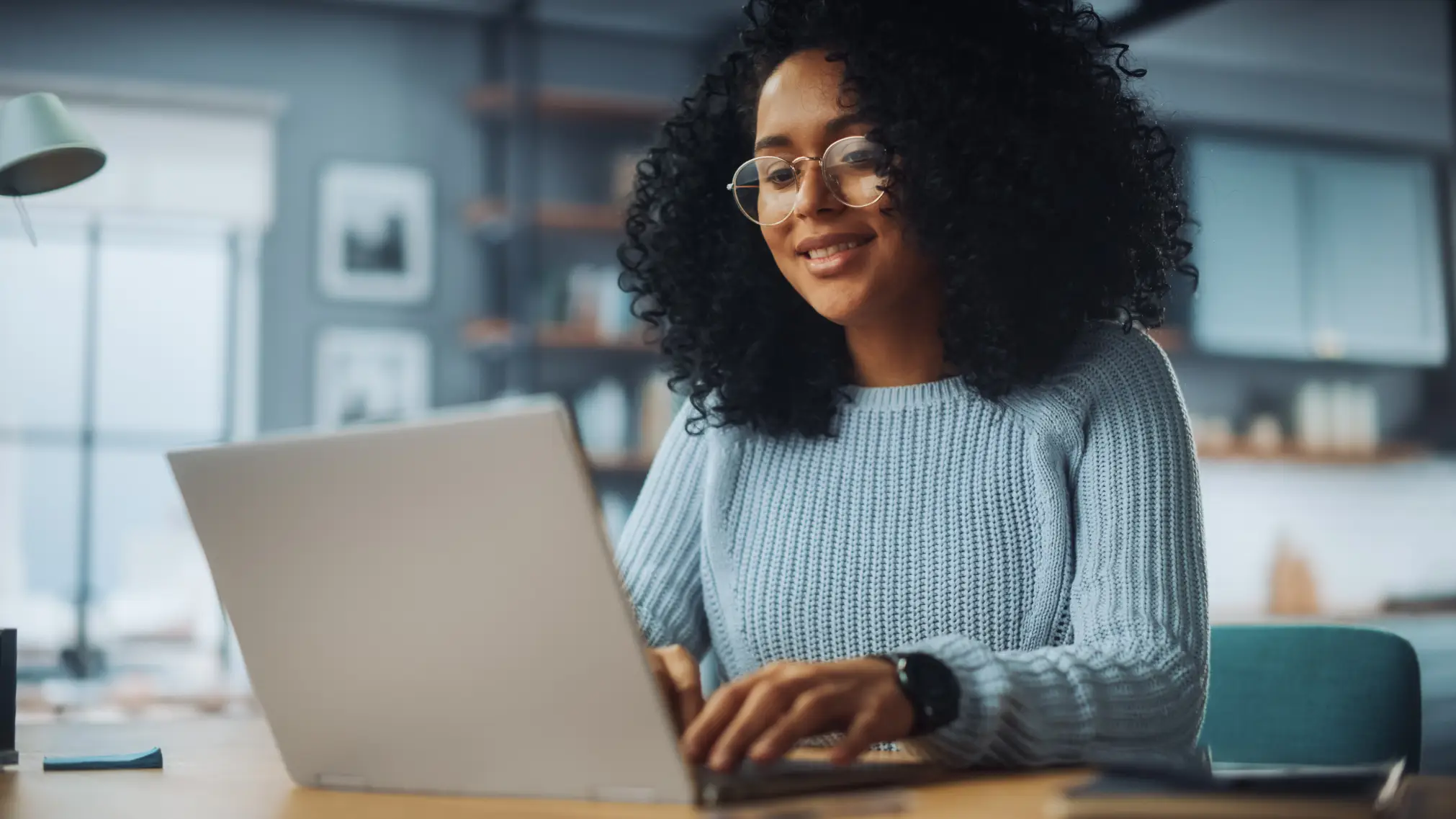 A woman with black curly hair wearing a light blue sweater and round glasses sits at a desk and types on her laptop.