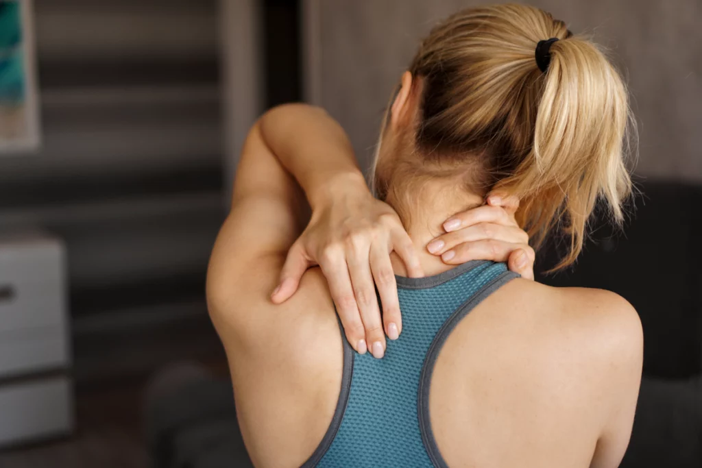 Back view of a blonde woman wearing a blue sports bra rubbing her neck and upper back in pain.