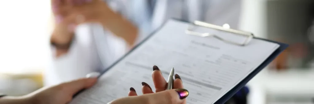 A close-up image of a form on a blue clipboard being held by hands with chrome-painted nails and a pen in the right hand.