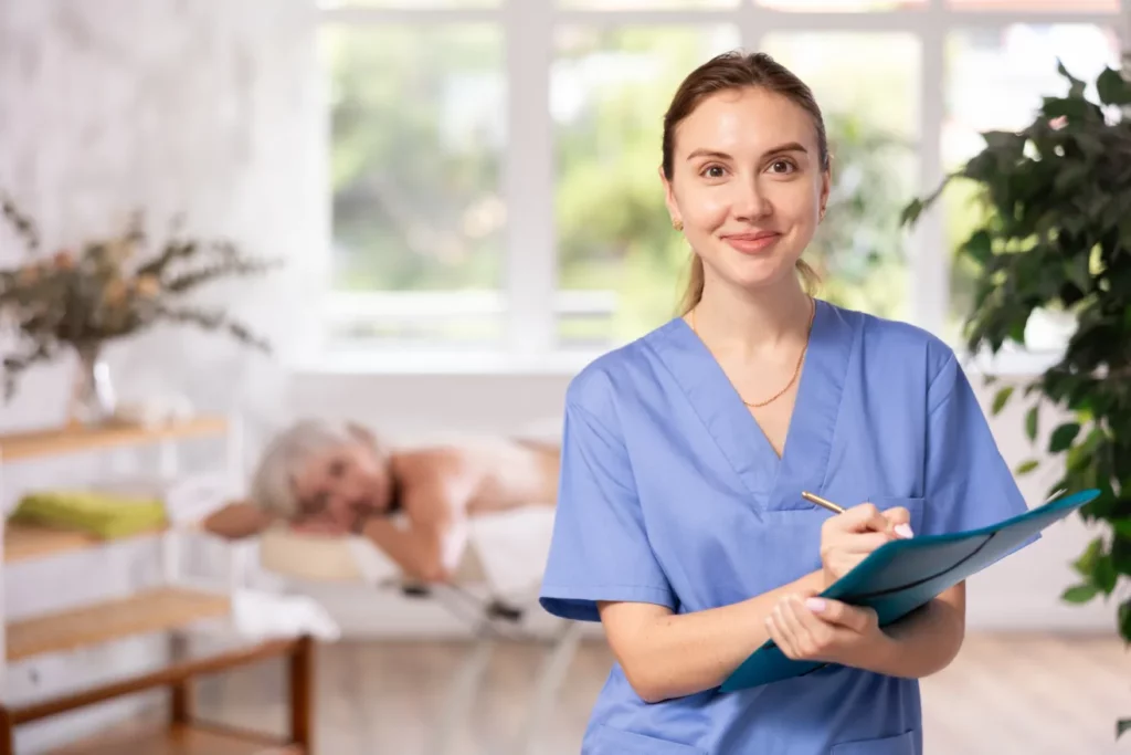 A female massage therapist wearing blue scrubs, smiles and writes on a blue folder in a massage room with a client lying on a massage table behind her.