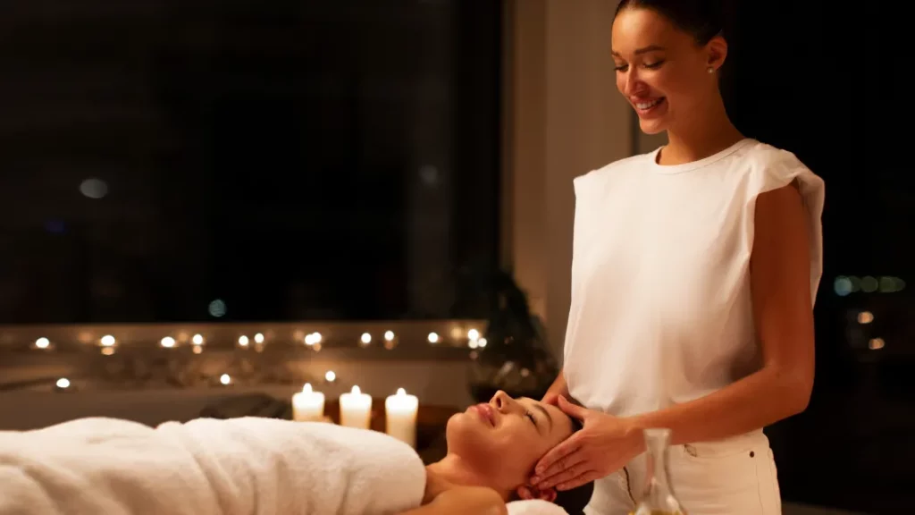 A massage therapist wearing a white top and pants performs a head massage on a client in a candlelit massage room.