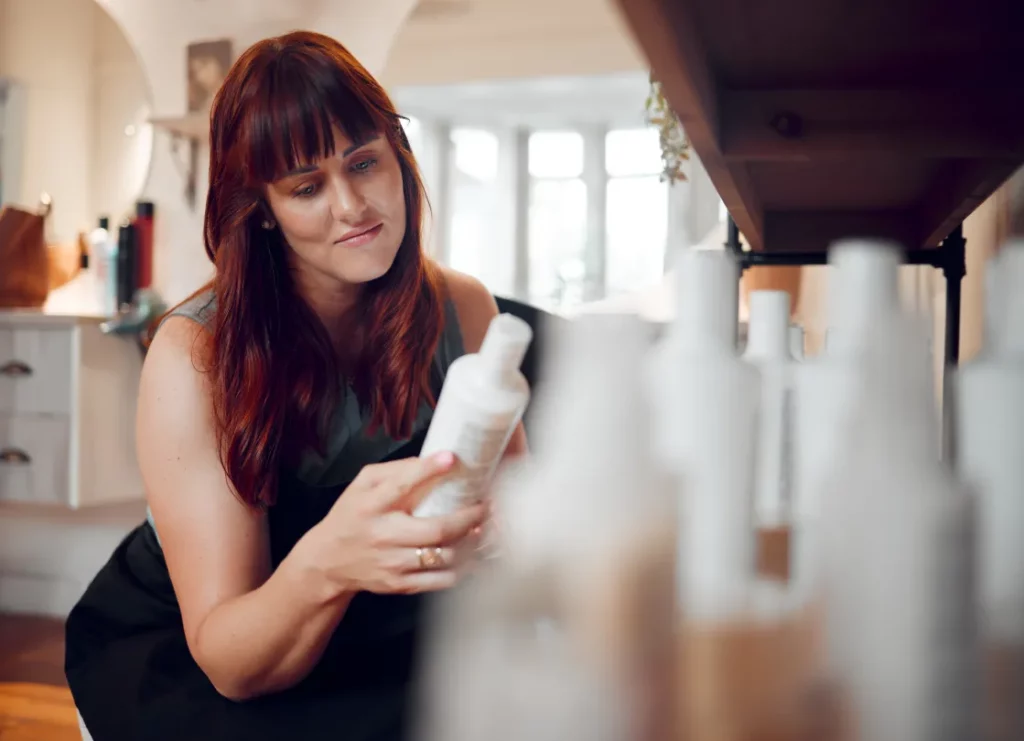 A salon owner with red hair checks a white product bottle in a salon.