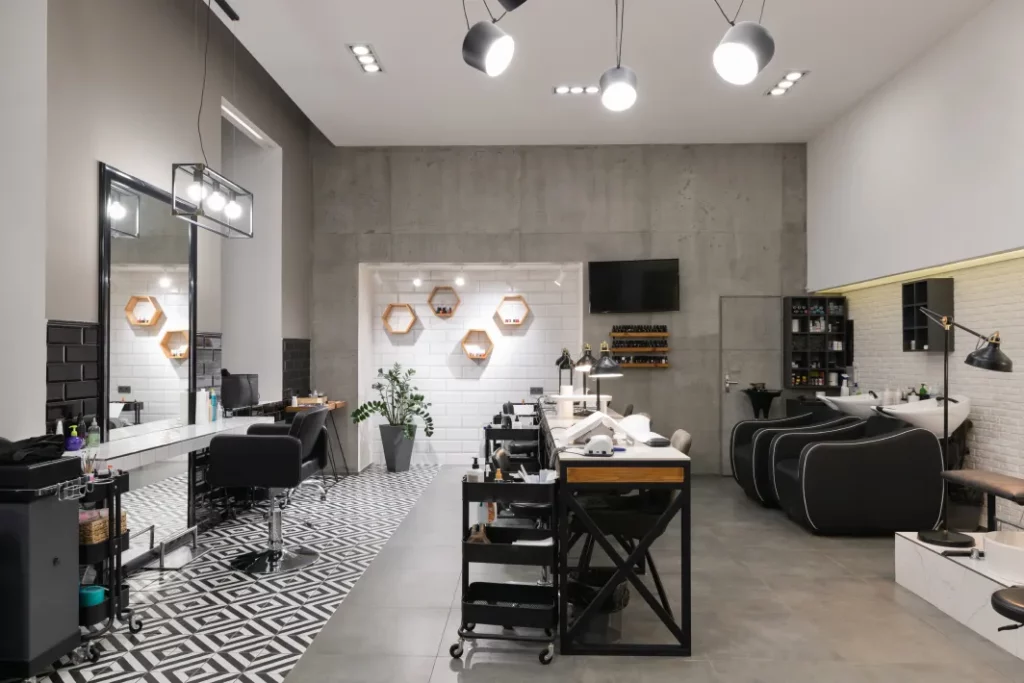 A modern salon in gray, white, and black tones with a mirrored station on the left, a long table in the middle, and several shampoo stations on the right. There's lighting hanging throughout and a white-and-black geometric tile on the left side of the floor.