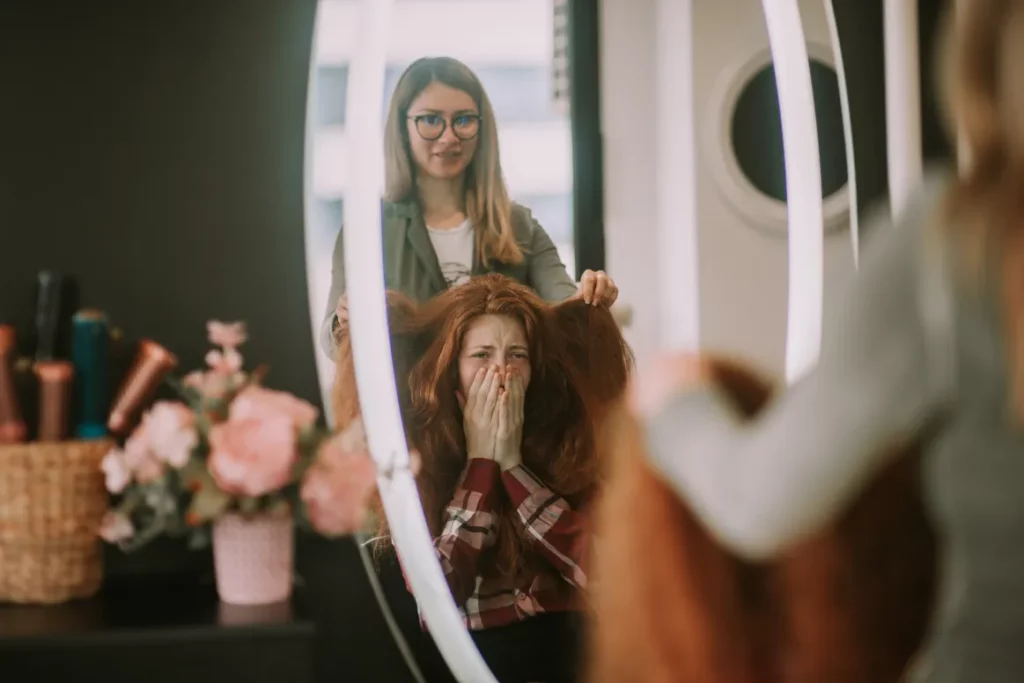 A client with long red hair covers her face in shock while a stylist touches the strands as they both look into the station mirror at a salon.