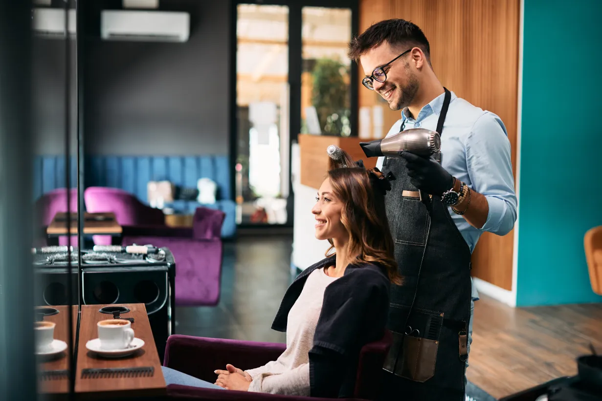 A hair stylist wearing a blue button-up shirt and a denim apron blow-dries a client's hair using a round brush in a salon with gray, brown, and teal walls.
