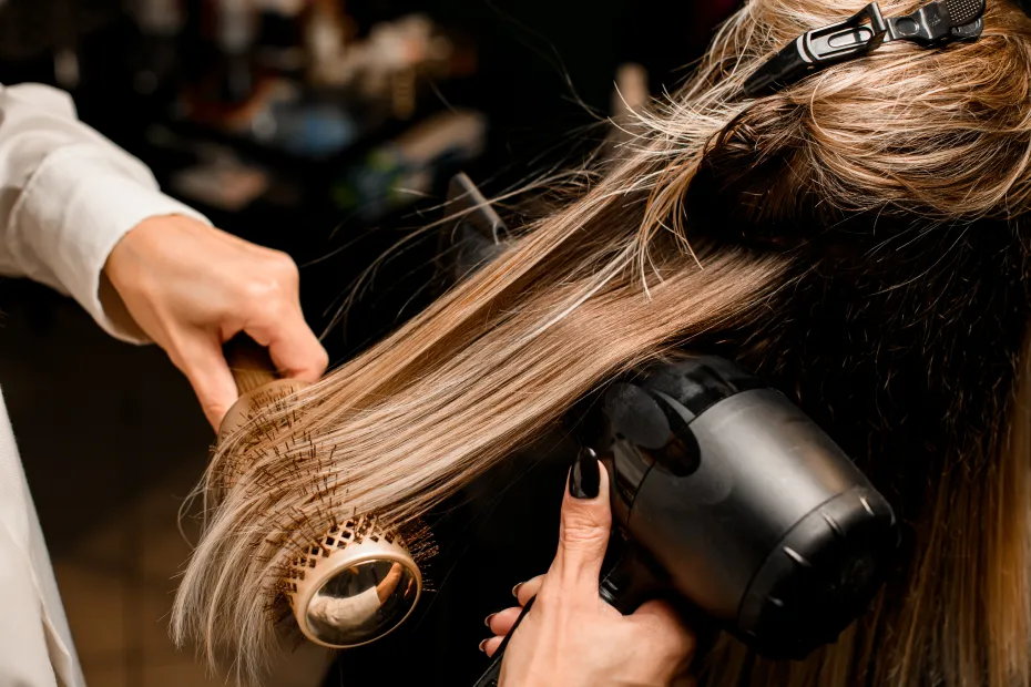 A stylist blow dries a client's light brown hair using a hair dryer and round brush.
