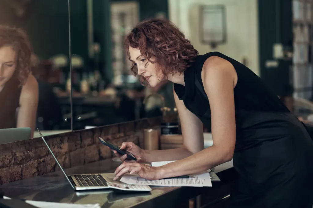 A stylist with short curly hair wearing a black top leans over a table while typing at a laptop and holding a smartphone.