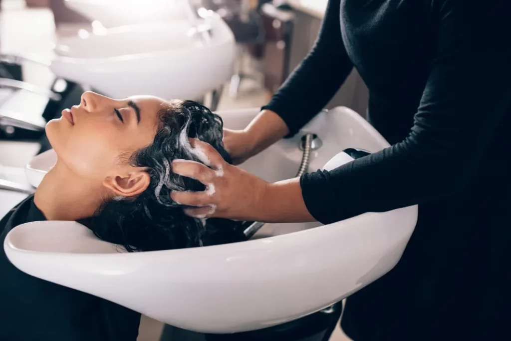 A client with dark brown hair receives a shampoo at a shampoo station from a stylist wearing a black long-sleeve top.
