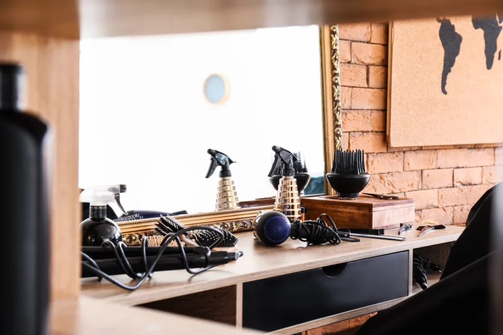 A salon station with a mirror, wooden shelving, spray bottle, hair dryer, flat iron and other tools against a red brick wall with wall art of a world map in the background.