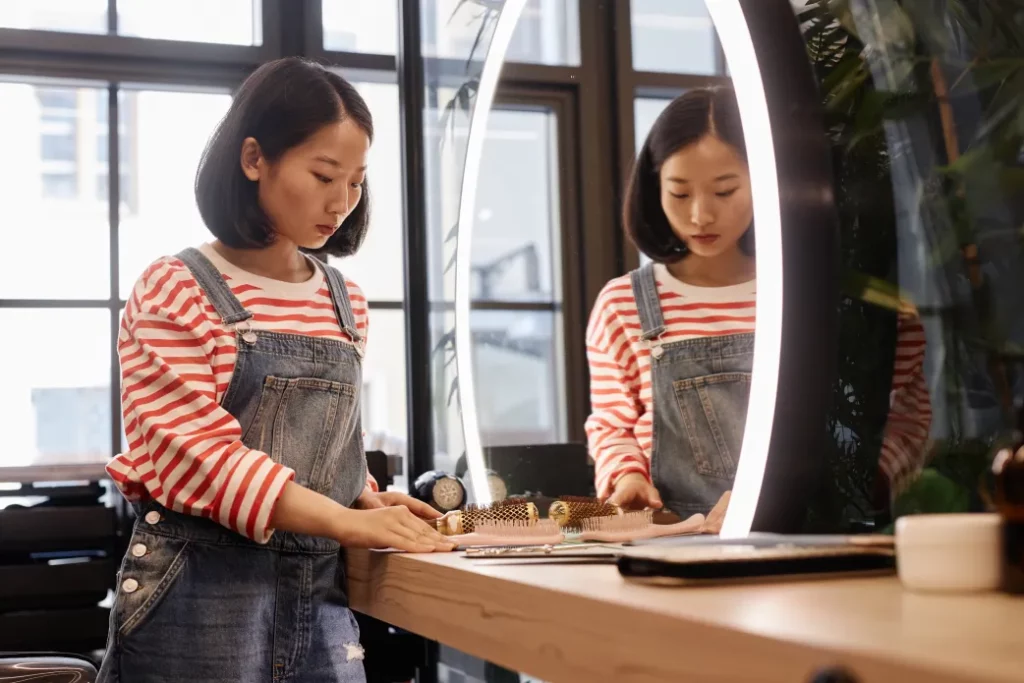 A stylist wearing a red-and-white stripe top and denim overalls looks over business papers at a salon station with a round lit mirror.
