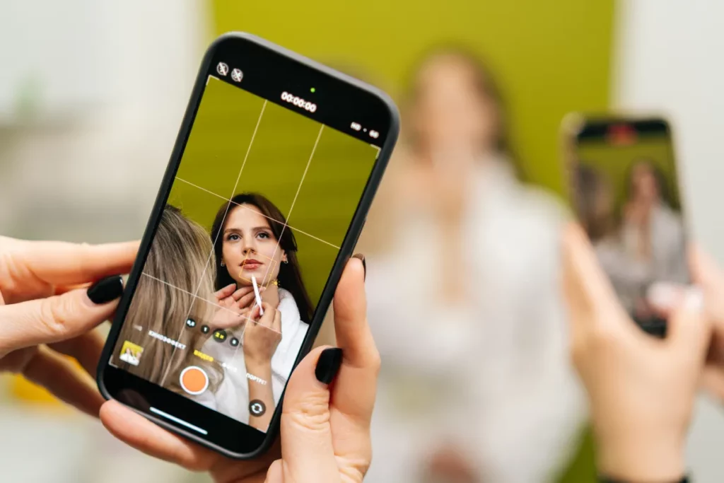 A close-up image of two hands holding a smartphone recording a makeup artist applying lip liner to a client against a green backdrop.