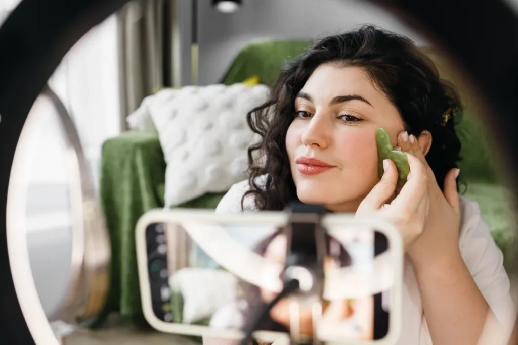 A massage therapist records herself massaging her face with a green gua sha tool in in front of a green sofa. The smartphone recording her is centered in a ring light in the foreground.
