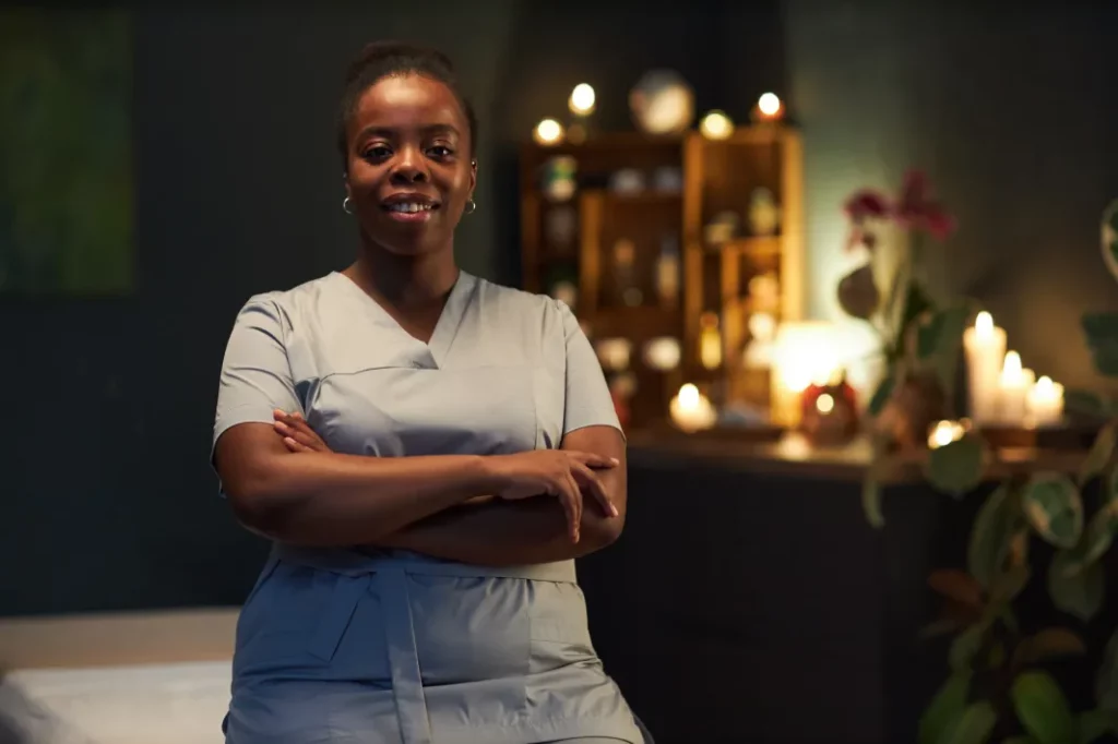 A massage therapist wearing blue scrubs smiles and crosses her arms in a candlelit massage room with a green wall, massage products, and plant decor in the background.