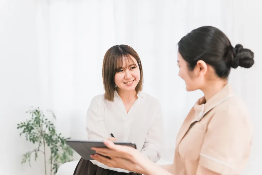A spa owner and a massage therapist review contracts on a business tablet in a white massage room with a green plant in the background.