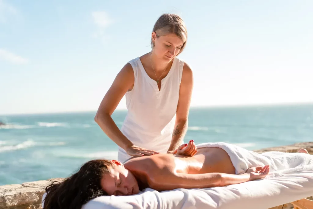 A massage therapist wearing a white tank top performs a massage on a client lying on a white massage table by a cliffside overlooking the ocean.