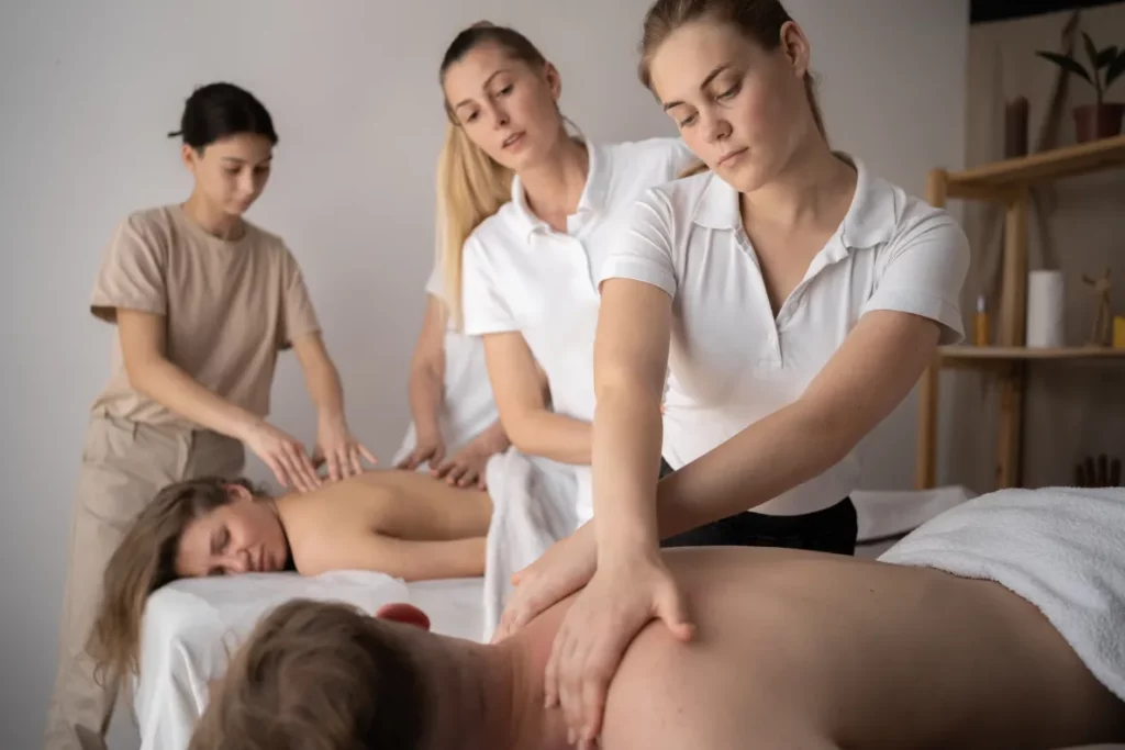 A group of massage therapists learn massage technique while working on clients lying down on massage tables in a massage room.