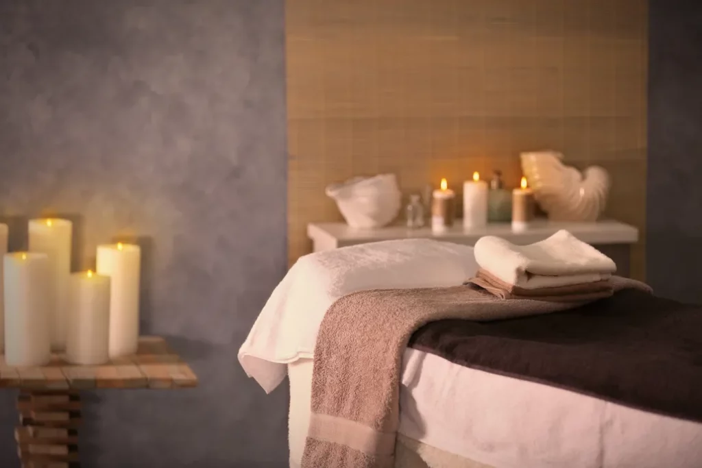A massage room set up with a massage table with white and neutral-toned towels, a table with white pillar candles, and a shelf with candles and decor in the background.