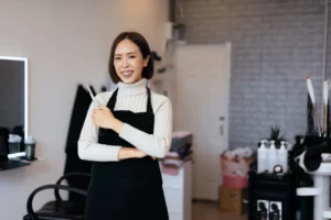 A salon owner wearing a white turtleneck top and a black apron smiles and holds a pair of scissors in her hair salon.