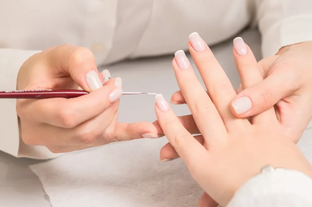 A nail technician holds a slim brush and paints a French manicure on a client's hands over a white paper towel.