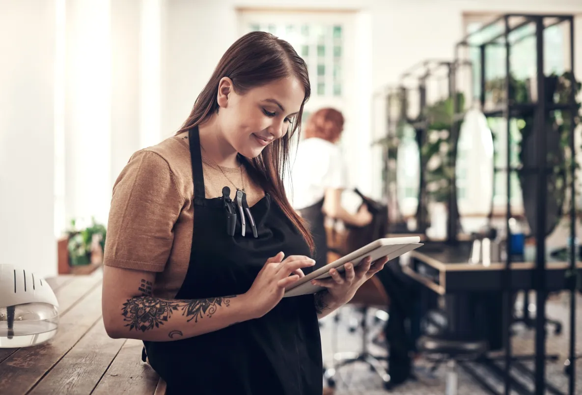 A salon owner wearing a brown shirt and a black apron smiles while tapping on a business tablet, performing a risk assessment in her hair salon with stations in the background.