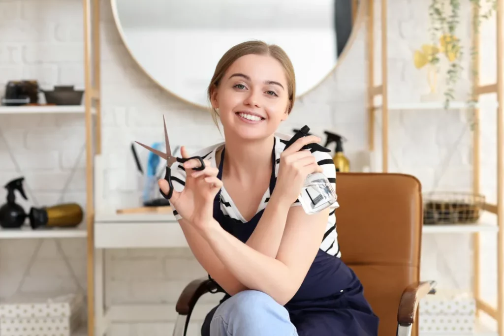 A hair stylist wearing a black-and-white striped top and a navy apron sits in a brown salon chair while holding scissors and a spray bottle.