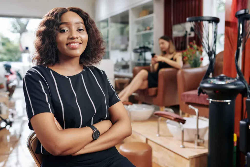 A nail salon owner wearing a black striped top smiles and poses in her nail salon with a client sitting in a chair and various sinks in the background.