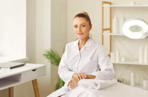 A female esthetician wearing a white button-up uniform smiles and leans against a white client bed in a studio with a white desk, a green plant, and white shelving behind her.
