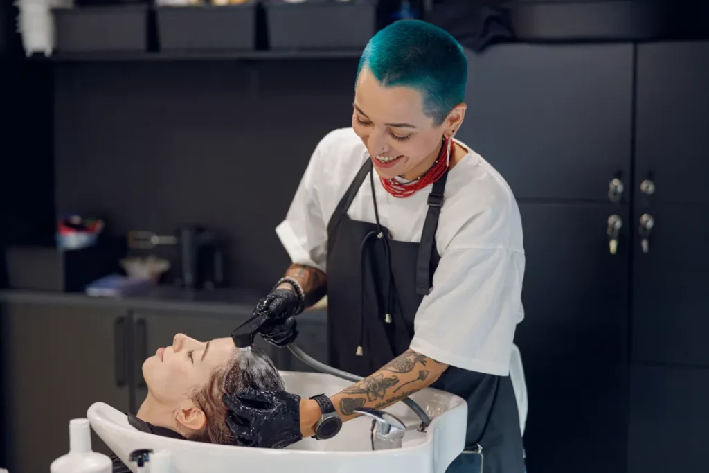 A hair stylist with blue hair wearing a white shirt and black apron washes a client's hair at a shampoo station in a salon.