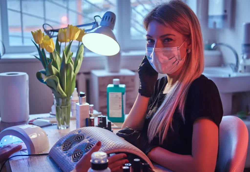 A nail technician wearing a face mask, a black shirt, and black gloves poses at a manicure desk with various nail equipment, yellow tulips in a vase, and an overhead lamp.