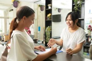 A salon owner prompts a new client to fill out a salon client intake form on a blue clipboard at the reception desk.