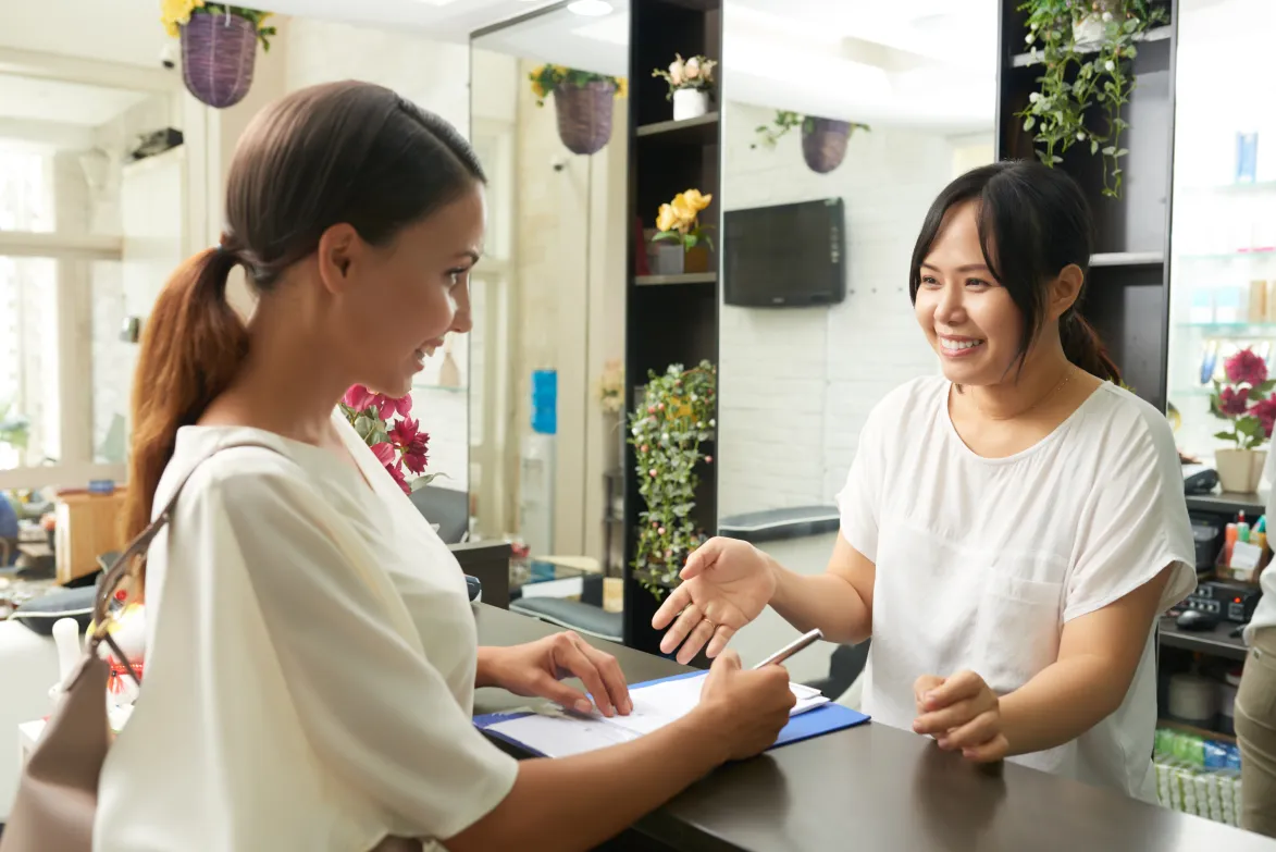 A salon owner prompts a new client to fill out a salon client intake form on a blue clipboard at the reception desk.