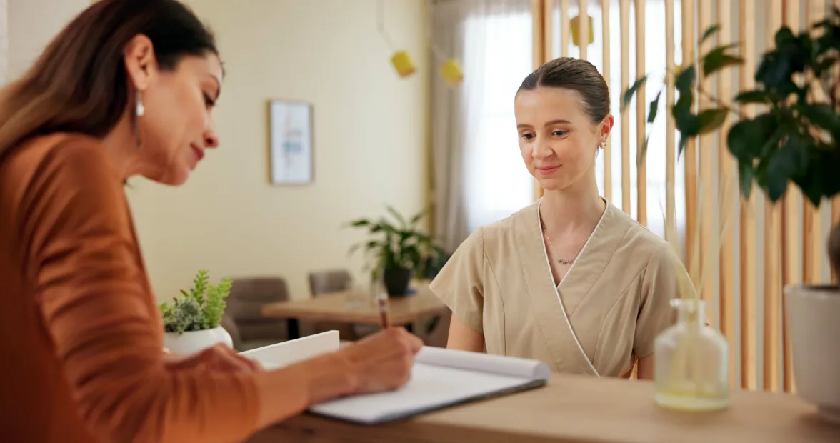 A salon receptionist wearing a beige wrap-around uniform watches a client sign a liability waiver on a clipboard at the reception desk of a salon decorated with green plants and soft-toned wood accents.