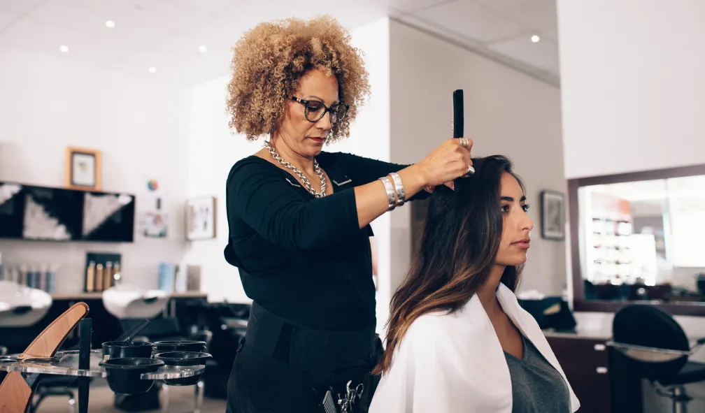 A hair stylist waring a long-sleeve black top, glasses, and a chunky silver necklace sections a client's hair with a comb at a salon station with shampoo stations and a large mirror in the background.