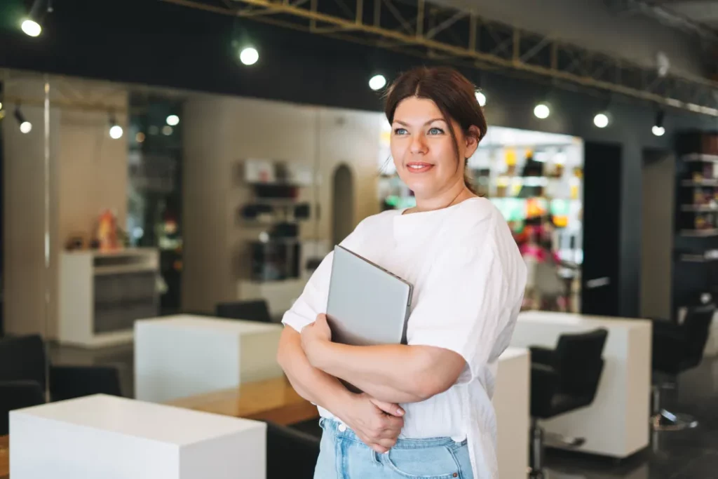 A salon owner wearing a white top and blue jeans smiles and holds a business laptop against her chest while standing in the reception area of a hair salon with large mirrors, white accents, and overhead lights in the background.