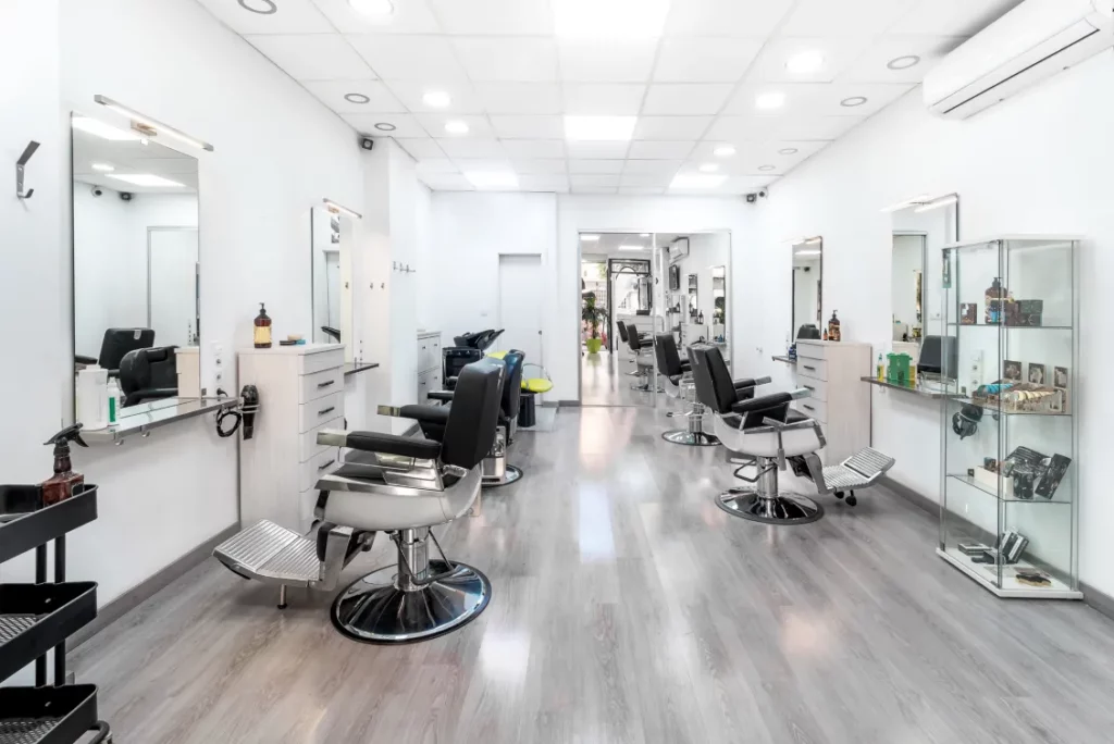 A brightly lit hair salon with rows of stations on either side, plus square mirrors, white cabinets, a glass product case, and a black tool tray.