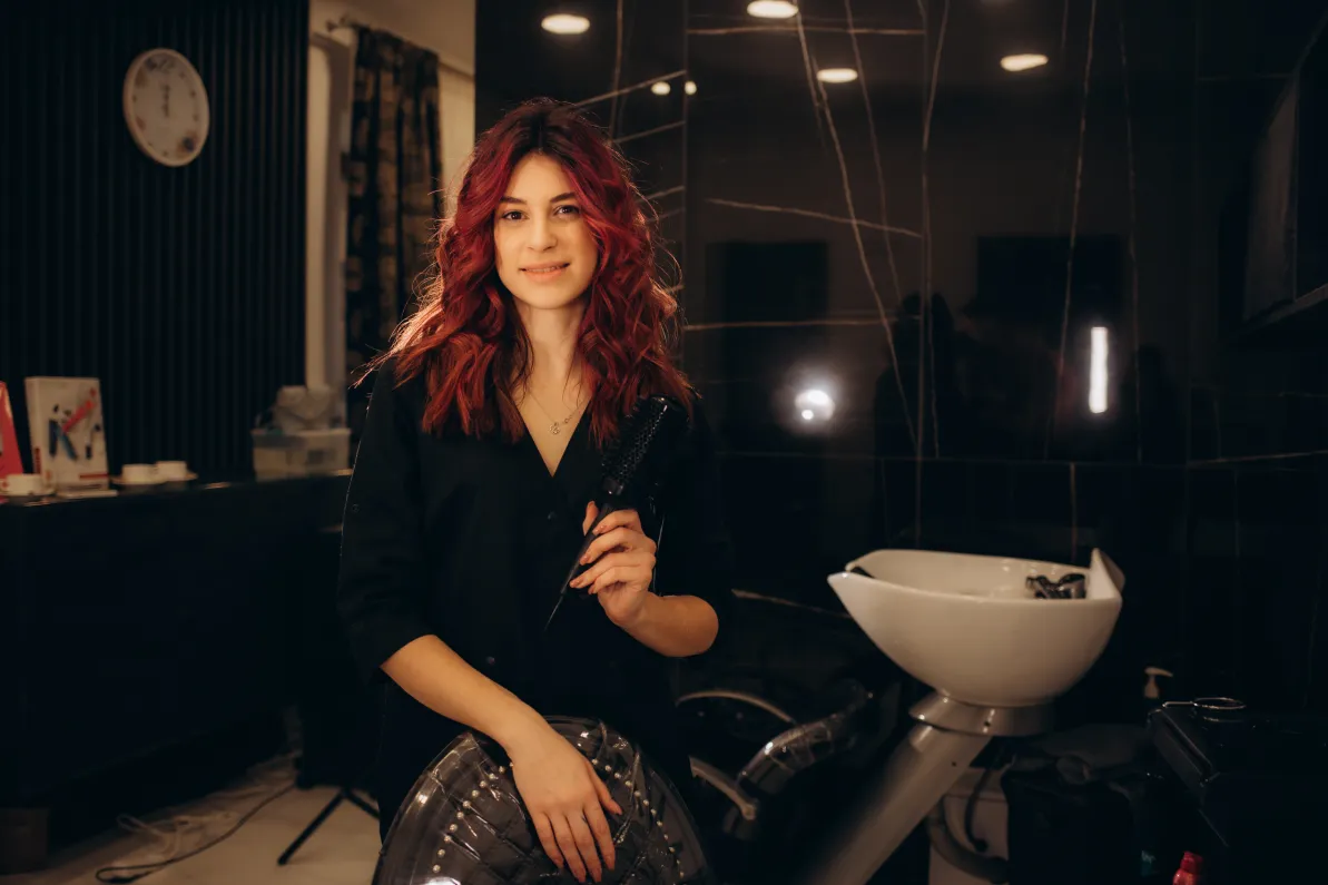 A salon owner with wavy red hair wearing a black top holds a hair tool while sitting in front of a shampoo station in a salon.