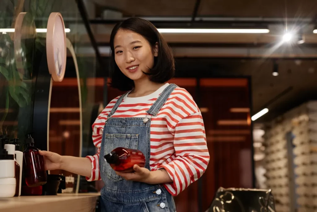 A salon owner wearing a red-and-white striped tee and denim overalls smiles and holds hair products in a salon.