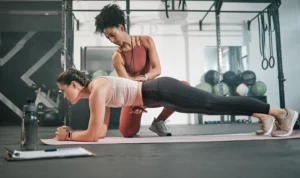 A personal trainer guides a client's elbow plank form in an industrial-style gym with a clipboard, water bottle in the foreground, and various workout gear in the background.