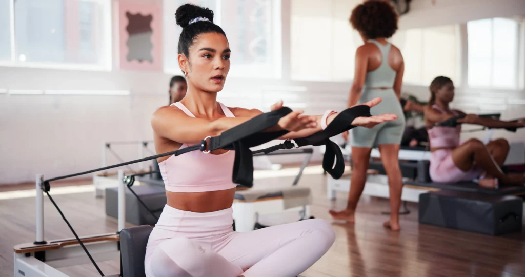 A woman wearing a pink workout set does arm exercises on a Pilates reformer machine in a Pilates studio.