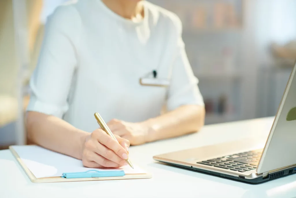 A massage therapist writes on a clipboard in front of a business laptop at a white desk.