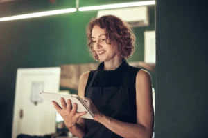A salon owner wearing a black sleeveless top and black apron smiles and taps on her business laptop in a salon with a green interior and overhead lights.