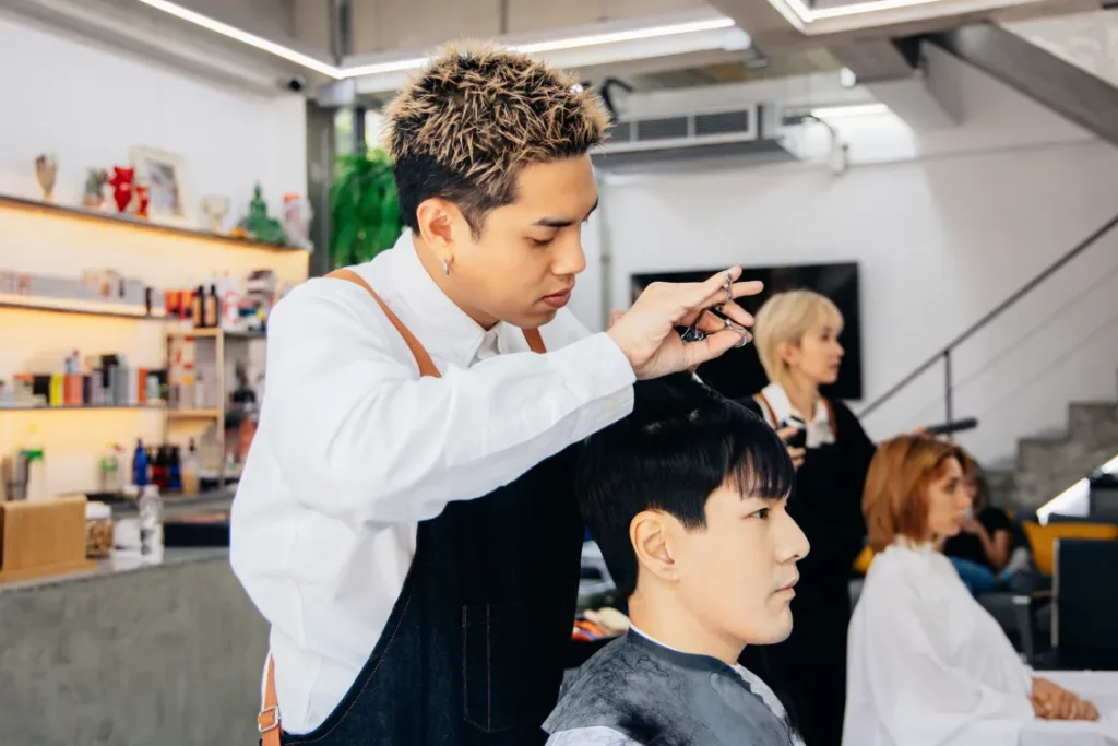 A hair stylist wearing a white long-sleeve button-up shirt and a navy apron cuts a client's hair with scissors in a salon.