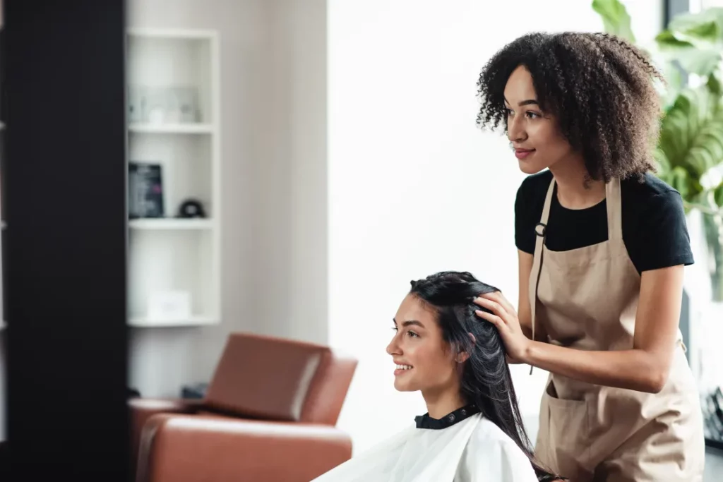 A hair stylist wearing a black shirt and a beige apron styles a client's hair at a salon station.