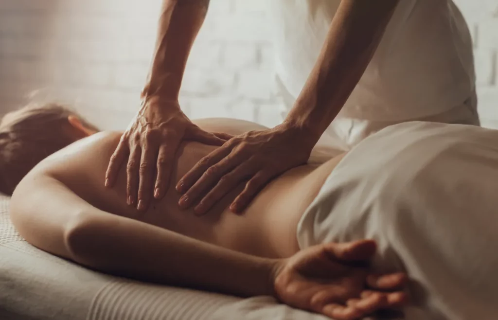 A massage therapist uses two hands to massage a client's back while the client lies on a white massage table in a white-brick massage room.
