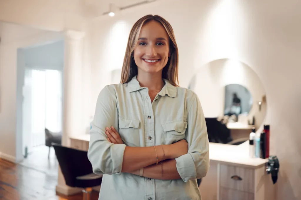 A salon owner wearing a mint-colored button-up shirt smiles and crosses her arms while standing in front of a salon station in a brightly lit salon with white walls and a circular mirror in the background.