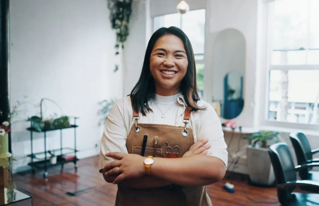 A salon owner wearing a white shirt and a brown apron with tools in its pockets smiles and crosses her arms while standing in a salon with multiple stations, open windows, green plants, and an oval mirror in the background.