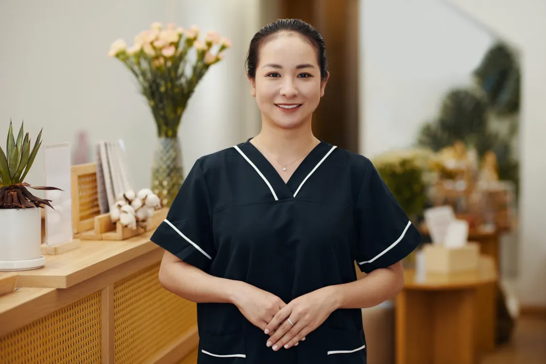 A spa owner wearing a black top with white trim smiles and clasps her hands in front of her as she stands in the reception area of a spa, which is decorated with warm wood tones and green plants.
