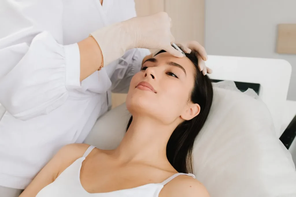 A medspa client wearing a white tank top lies back on a lined chair and receives an injectable in her forehead, administered by a medical aesthetician wearing a white coat and disposable gloves.