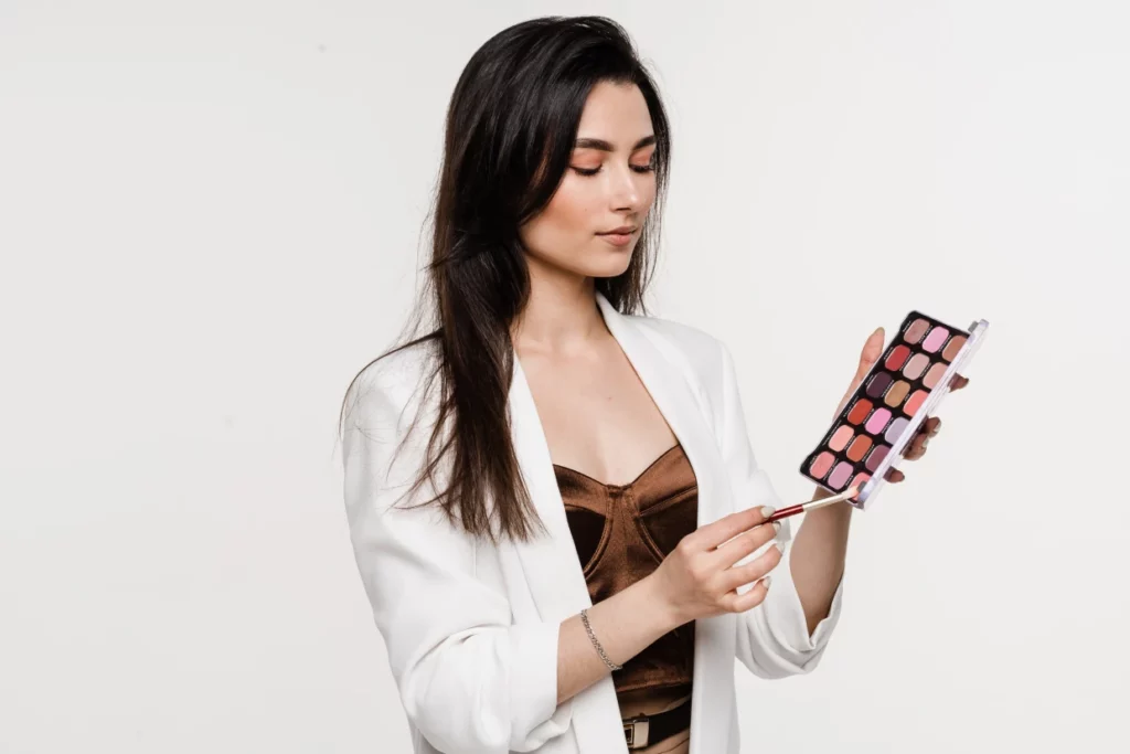 A makeup artist wearing a brown corset top and a white blazer presses her eyeshadow brush into a multi-pan eyeshadow palette in pink tones.